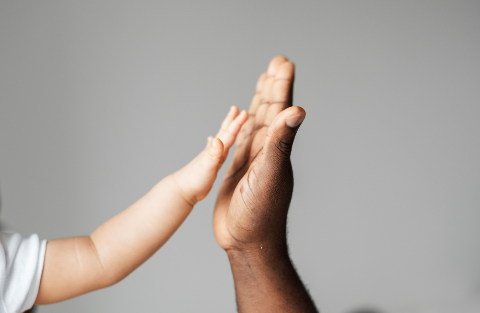 Imagem de um adulto e um bebê dando um “high five” com as mãos, simbolizando conexão, cuidado, aprendizado e parceria entre gerações, em fundo neutro