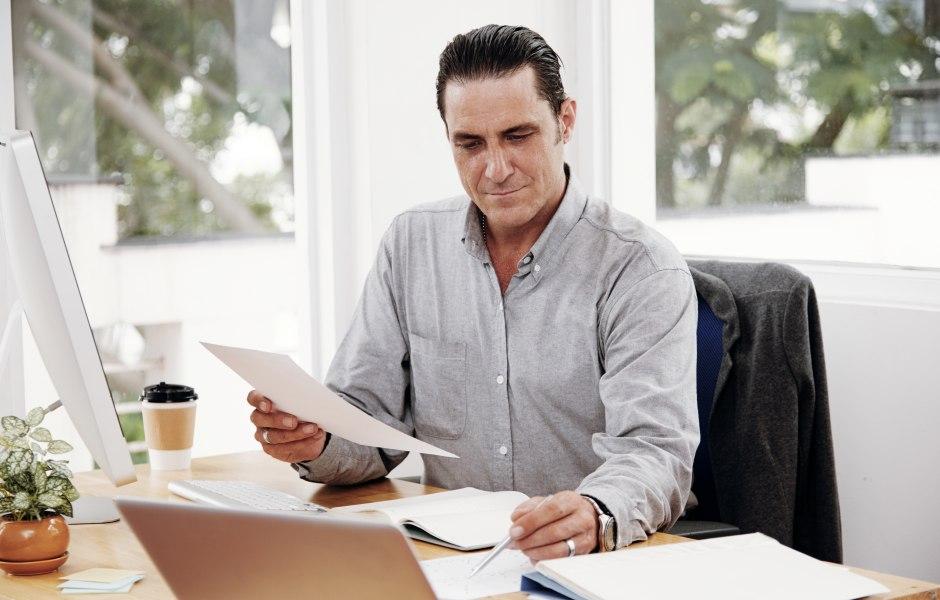 Homem de camisa social cinza trabalhando concentrado em sua mesa de escritório iluminada. Ele analisa um documento em papel enquanto segura uma caneta, cercado por um notebook, monitor e uma xícara de café.