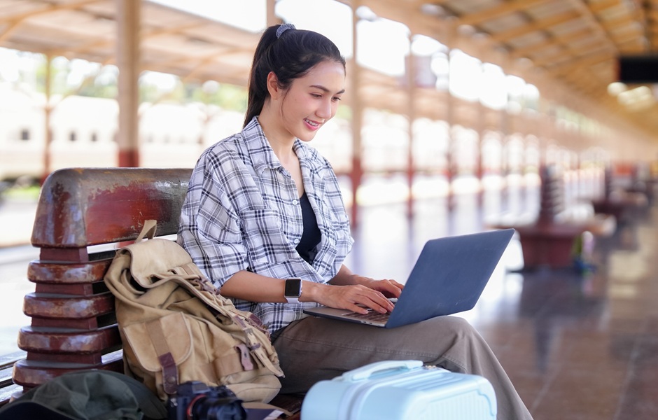 Jovem mulher usando laptop em estação de trem, sentada em banco com mochila e mala ao lado, aproveitando conexão Wi-Fi ao ar livre.