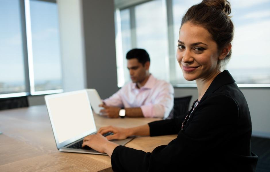 Mulher sorridente com cabelo preso em um coque digita em um notebook em uma mesa de escritório. Ao fundo, um homem desfocado trabalha segurando um tablet.