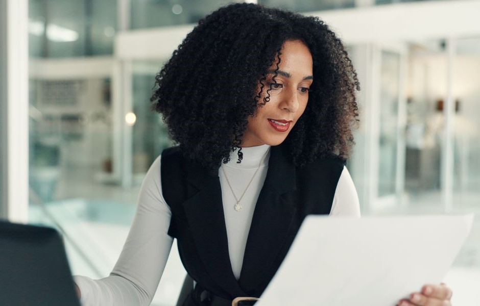 Mulher negra com cabelo cacheado lendo um documento na rotina diária de trabalho, vestindo roupas elegantes e com expressão focada, em ambiente moderno.