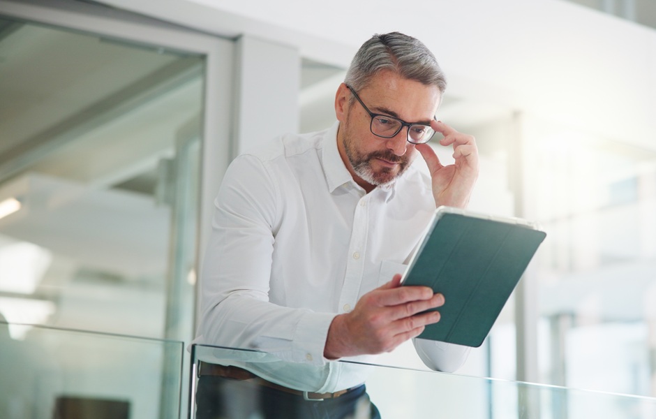 Homem de meia-idade usando óculos e tablet, com expressão de concentração, em ambiente corporativo moderno, representando inovação e tecnologia.