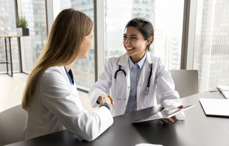 Duas médicas felizes em uma clínica moderna trocando um aperto de mãos, simbolizando confiança e parceria no cuidado à saúde, com fundo de janelas com vista panorâmica da cidade.