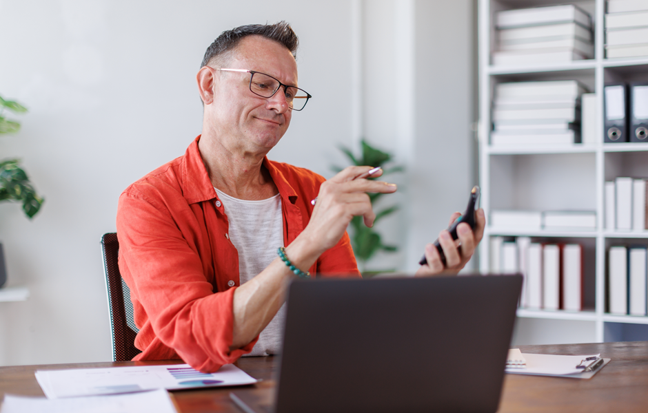 Homem sorridente usando smartphone em escritório moderno, com estante de livros ao fundo e papel na mesa.