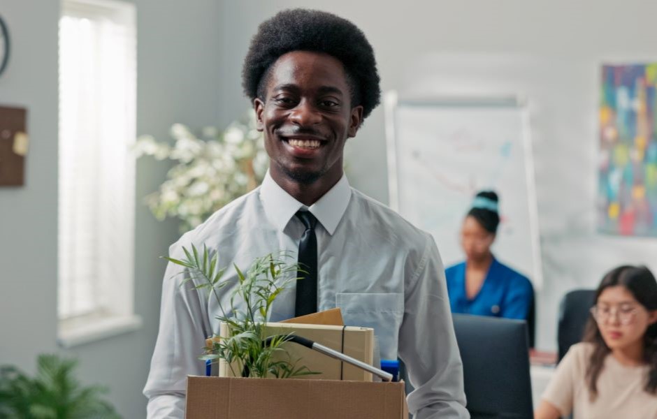 Homem sorridente e profissional em ambiente de escritório, segurando caixas com objetos e plantas, representando integração e crescimento profissional.
