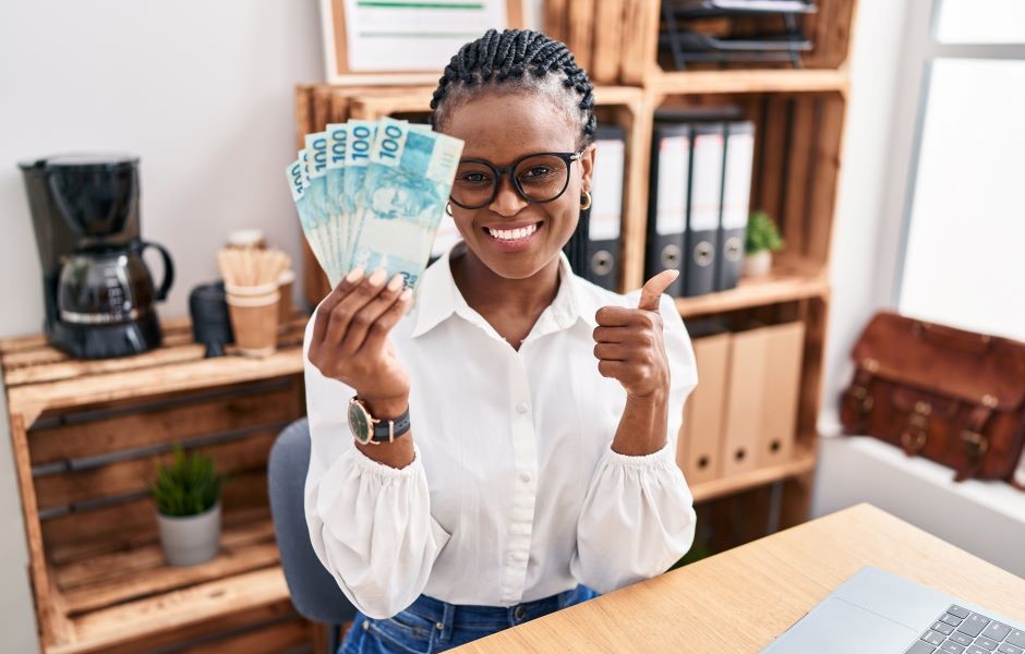 Mulher sorridente segurando dinheiro de 100 reais e fazendo um gesto de positivo, representando sucesso financeiro.