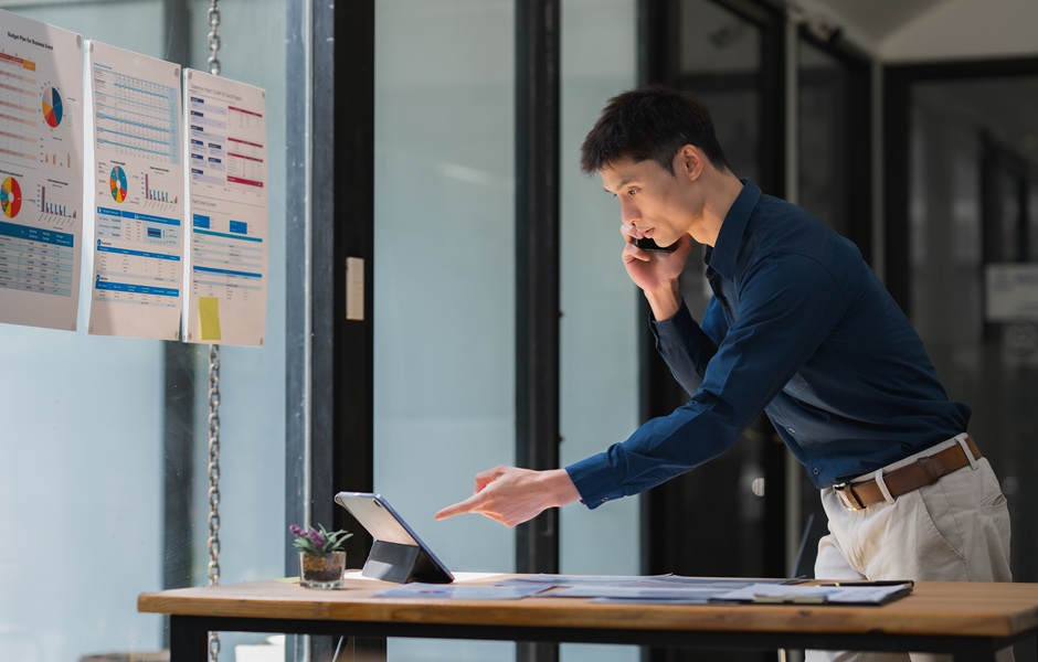 Homem de negócios falando ao telefone enquanto trabalha em uma mesa com tablet e documentos, no ambiente de escritório moderno.