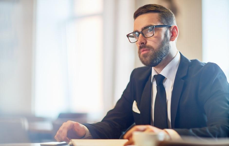 Homem de barba e óculos usando terno e gravata, sentado à mesa com um semblante sério e focado enquanto olha para o lado.