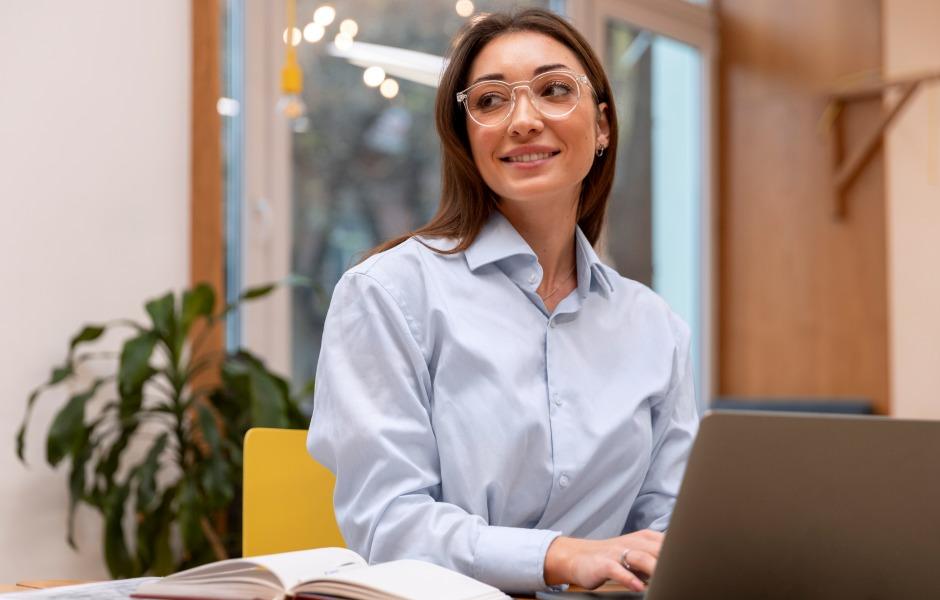 Mulher jovem de óculos e camisa social azul sorri enquanto trabalha em um notebook em um ambiente de escritório.