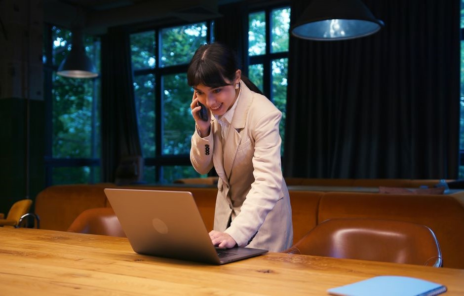 Mulher de negócios sorridente falando ao telefone enquanto trabalha em seu laptop em uma sala moderna com janelas grandes e vista para a natureza.