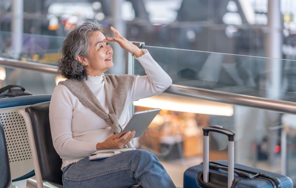 Mulher de meia idade sorrindo e segurando um tablet, sentada em aeroporto com mala de viagem ao lado, buscando informações de viagem ou aeroporto.