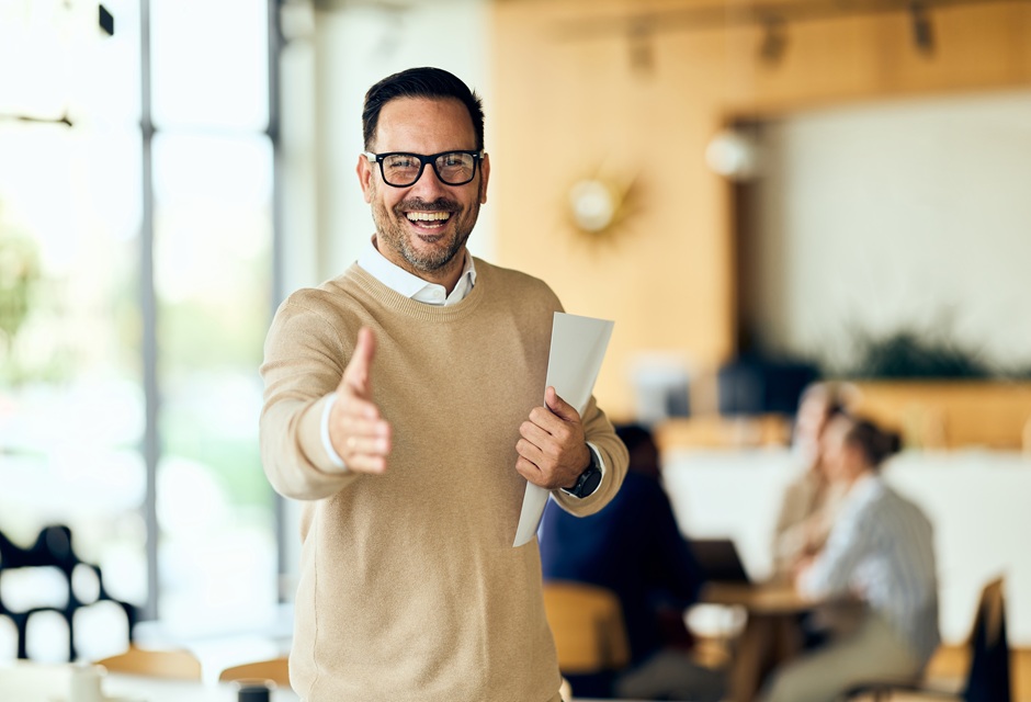 Homem sorridente fazendo gesto de aprovação com o dedo indicador para a câmera, em ambiente de escritório moderno, transmitindo positividade e aprovação.