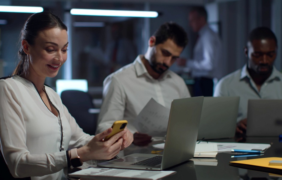 Equipe de profissionais trabalhando em um escritório moderno durante a noite, com foco em uma mulher sorridente usando o celular.