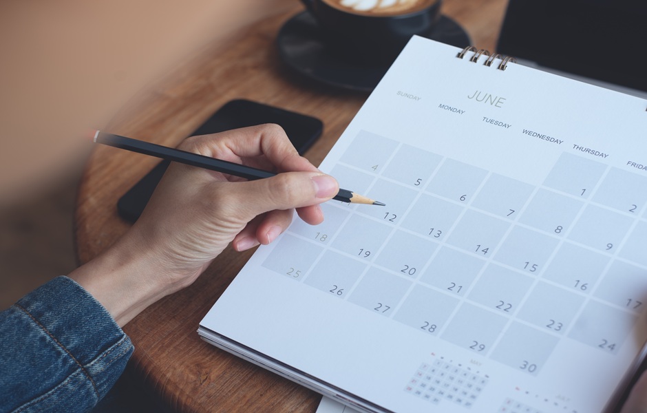 Pessoa anotando algo em um calendário de junho em uma mesa de madeira, com celular e copo ao fundo, lembrando planejamento de rotina ou agenda.
