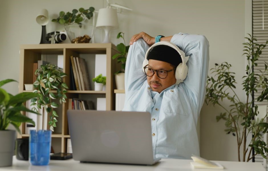 Homem de óculos e fones de ouvido descansando em um ambiente de escritório, sentado na frente de um laptop, rodeado por plantas e livros, sugerindo relaxamento no trabalho.