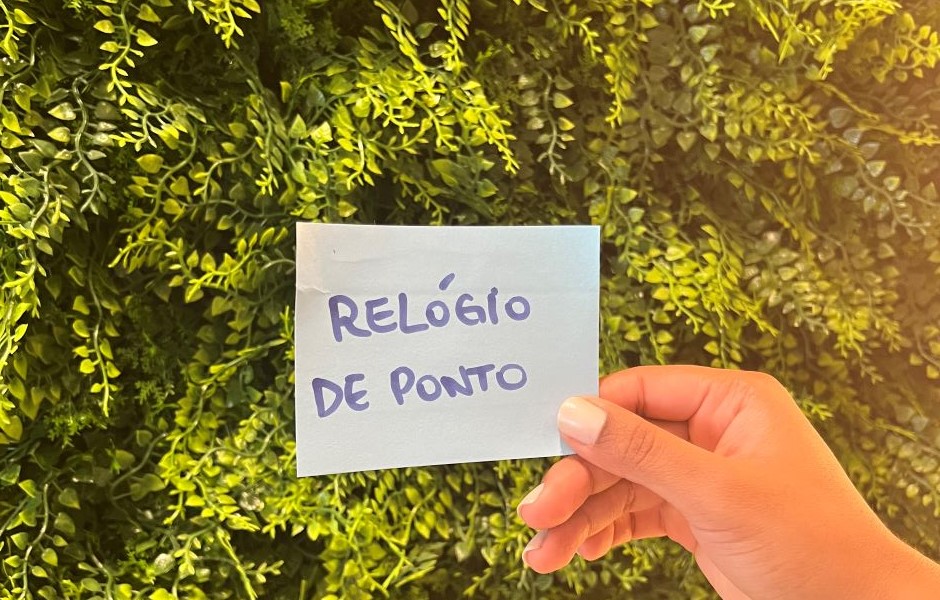 Mão segurando um papel com a frase 'Relógio de Ponto' em frente a uma parede de plantas verdes, destacando o tema de controle de ponto e registros de presença.