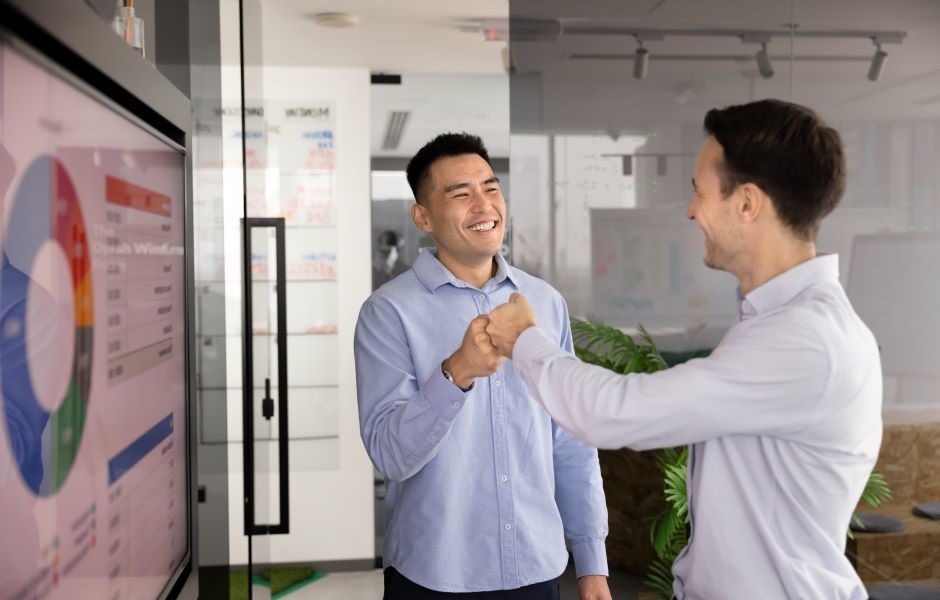 Dois homens de negócios sorridentes celebrando com um cumprimento amigável em um escritório moderno, destacando um ambiente de sucesso profissional.
