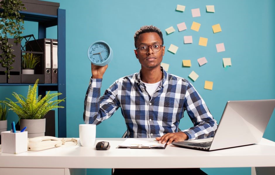 Homem jovem sentado em escritório com mesa de trabalho, segurando um relógio de parede em uma mão, representando gerenciamento de tempo e produtividade.