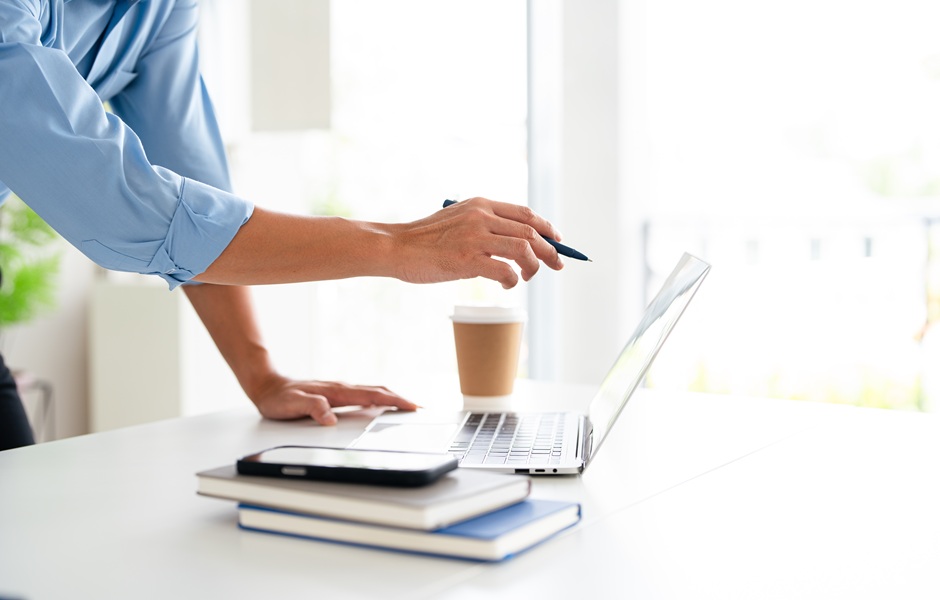 Pessoa usando laptop em ambiente de escritório com caneta na mão, acompanhada de café e cadernos, focada em trabalho ou estudo.